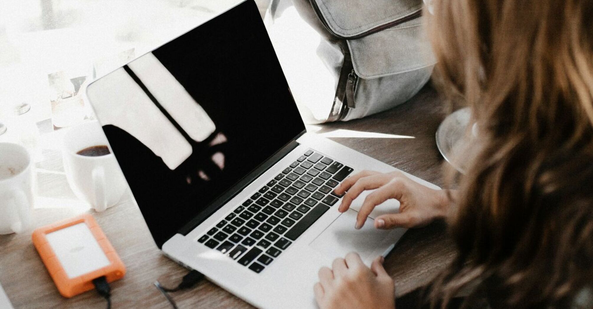 A woman working on a laptop near a window with a backpack on the table