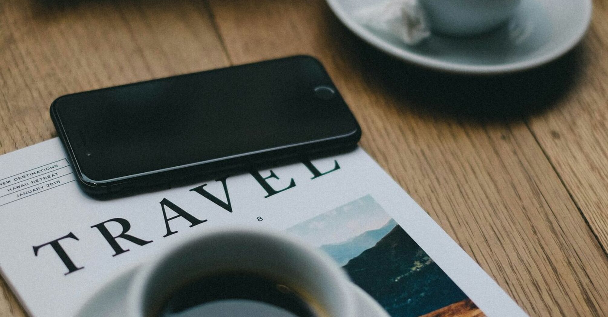 A magazine titled "TRAVEL" with two cups of coffee and a smartphone on a wooden table