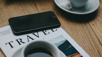 A magazine titled "TRAVEL" with two cups of coffee and a smartphone on a wooden table