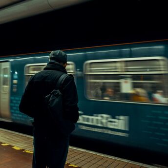 A man in a dark jacket and backpack stands on a train platform as a ScotRail train passes by in motion