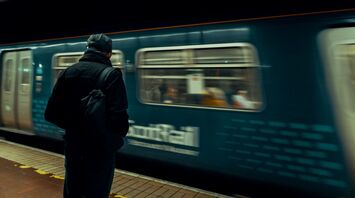 A man in a dark jacket and backpack stands on a train platform as a ScotRail train passes by in motion