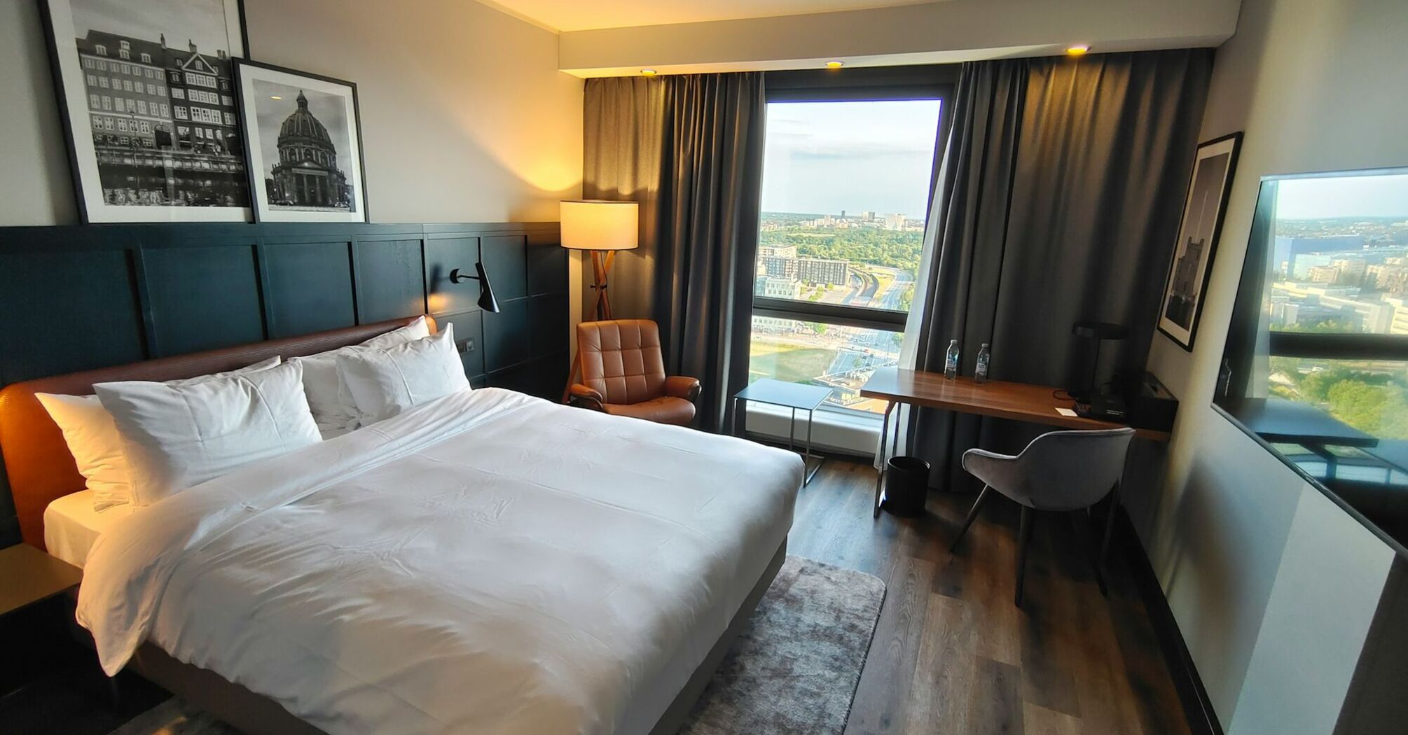 A modern hotel room with a large bed, stylish decor, and a city view through a floor-to-ceiling window