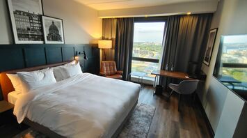 A modern hotel room with a large bed, stylish decor, and a city view through a floor-to-ceiling window