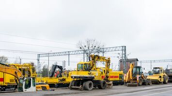 Yellow construction vehicles and equipment stationed near a railway, with overhead power lines and tracks in the background