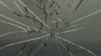 Shattered glass with a spiderweb pattern of cracks, reflecting light and a blurred outdoor scene in the background