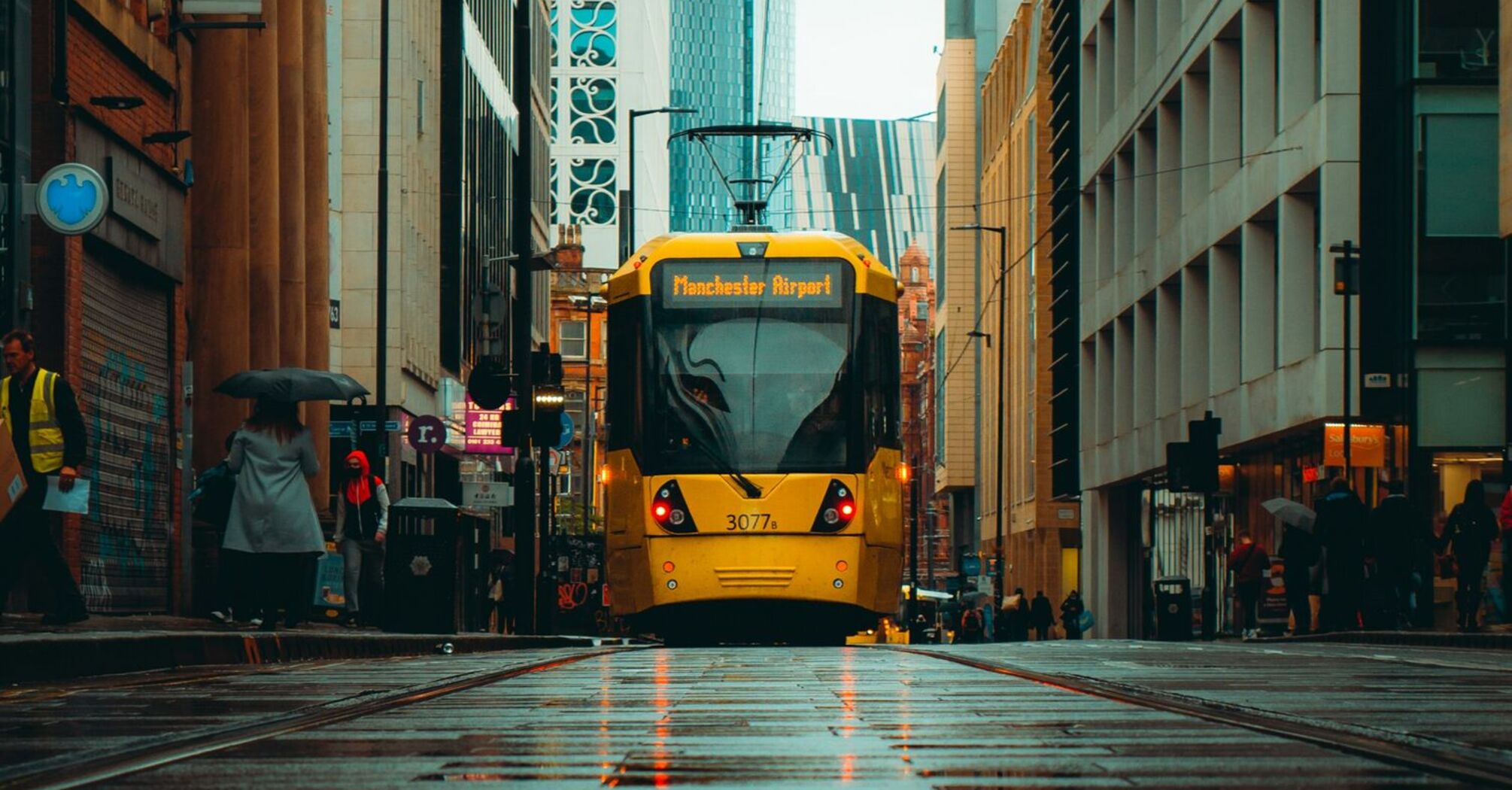 A yellow Manchester tram traveling through a wet city street with modern and historic buildings in the background