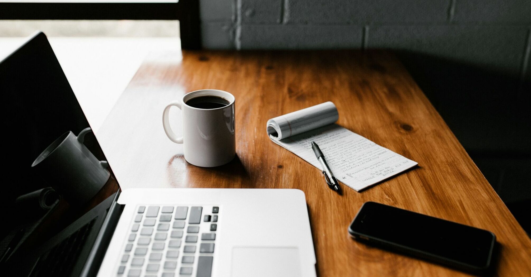 A workspace with a laptop, a cup of coffee, a notepad with handwritten notes, a pen, and a smartphone on a wooden desk near a window