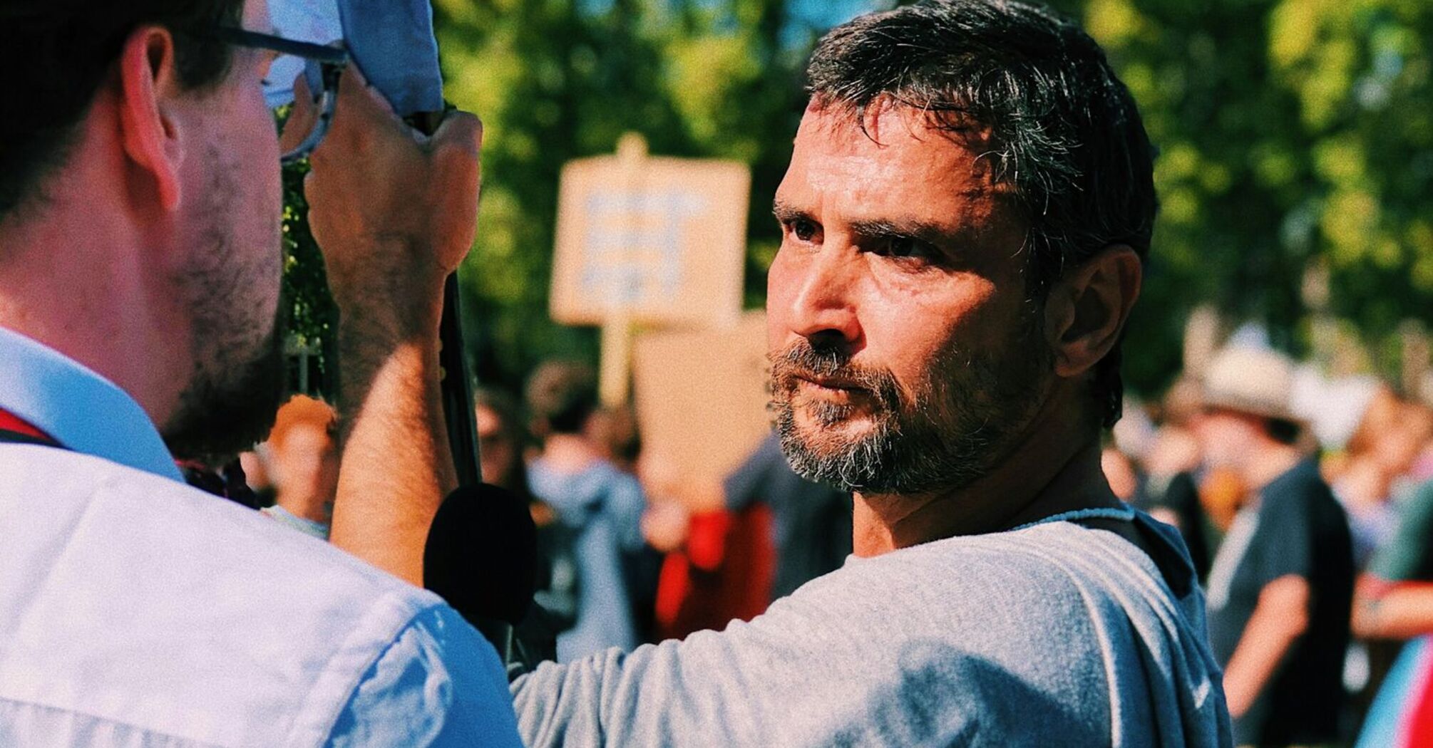 A man at a protest holds a sign while engaging in a discussion, with a crowd in the background