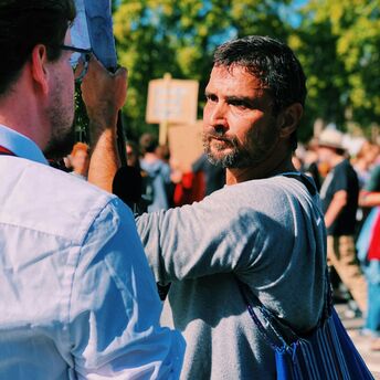 A man at a protest holds a sign while engaging in a discussion, with a crowd in the background