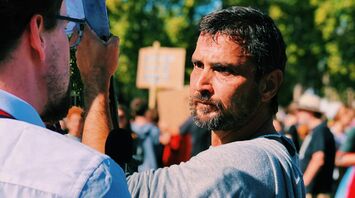 A man at a protest holds a sign while engaging in a discussion, with a crowd in the background