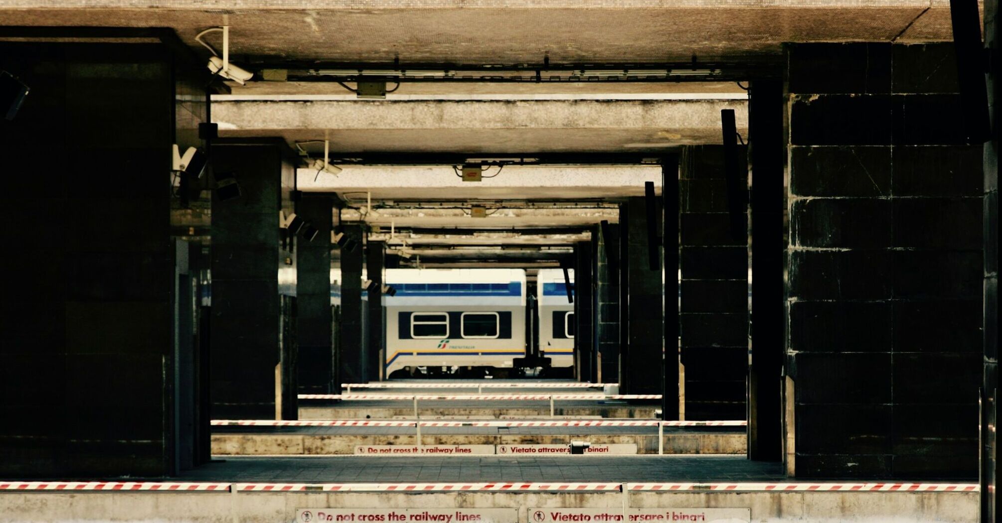 A quiet railway platform in Italy with a train in the background, marked with warning signs prohibiting crossing the tracks