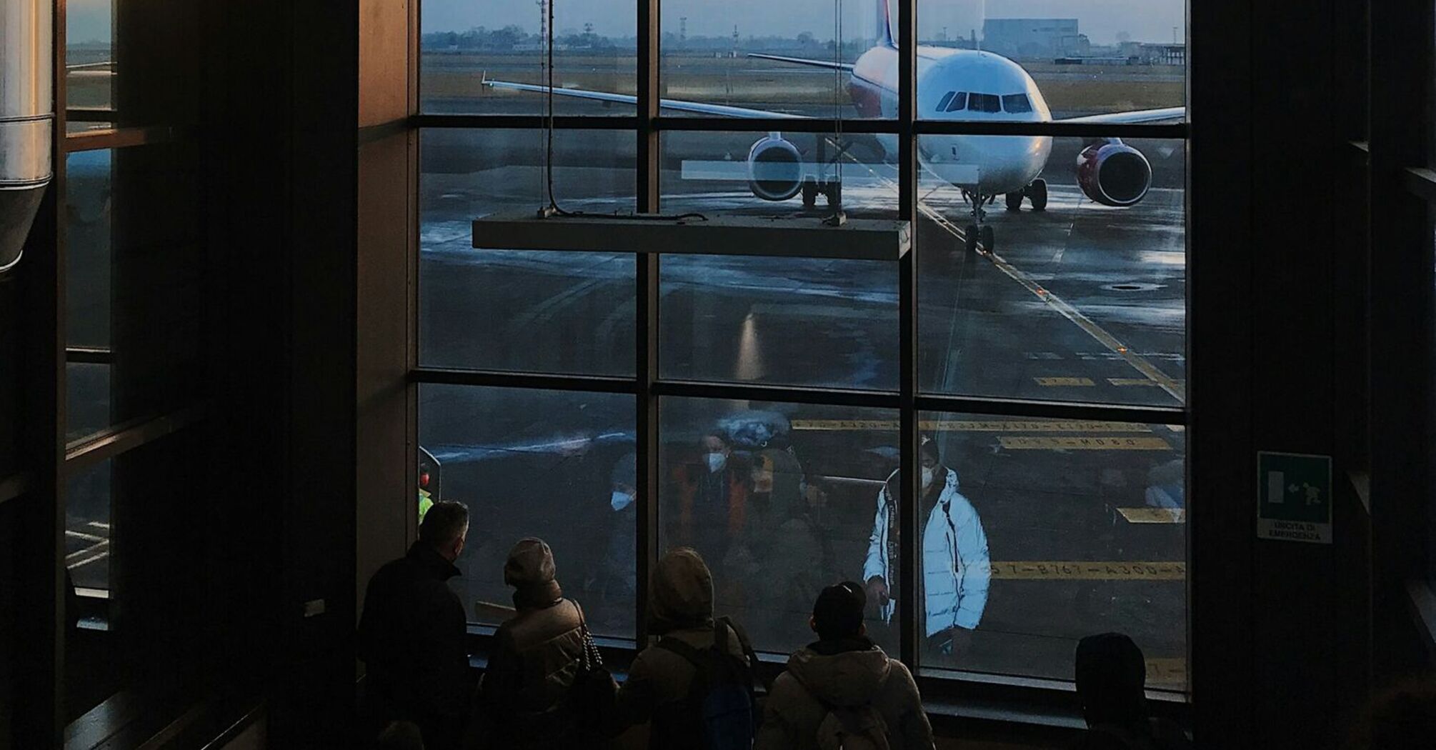 Passengers at an Italian airport terminal watching a parked airplane through large windows during sunset