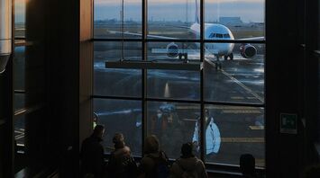 Passengers at an Italian airport terminal watching a parked airplane through large windows during sunset