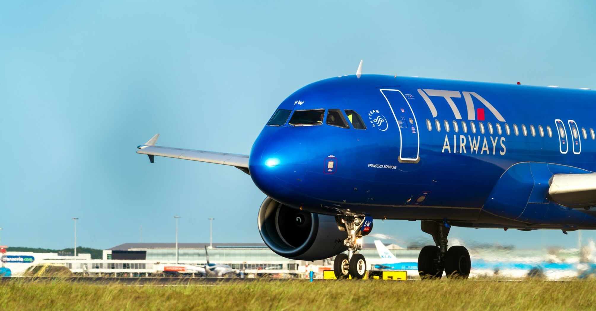 A blue ITA Airways Airbus A320 taxiing on the runway with an airport terminal in the background