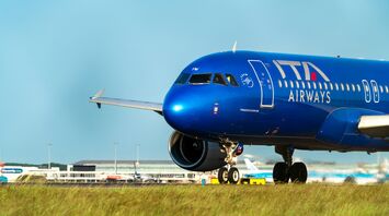 A blue ITA Airways Airbus A320 taxiing on the runway with an airport terminal in the background
