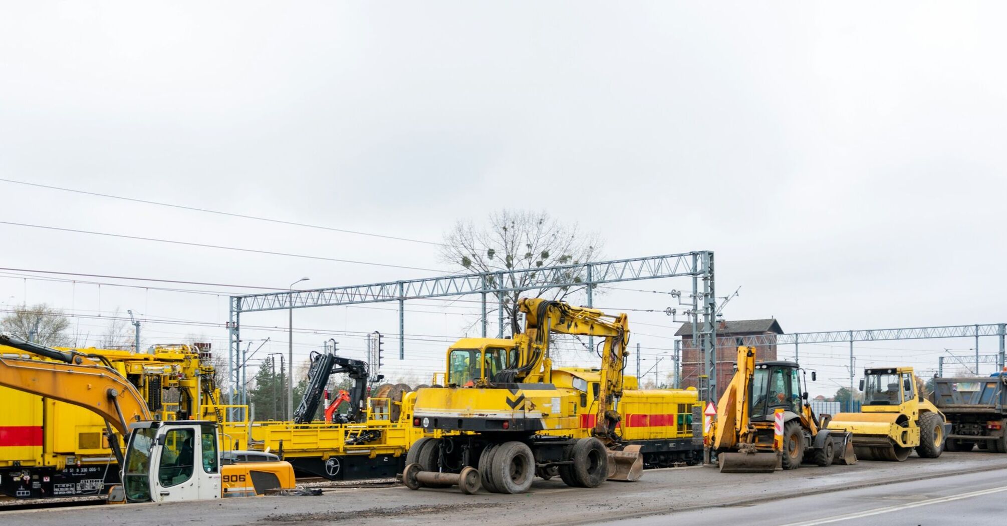 Heavy construction equipment at a railway site undergoing maintenance and upgrades, with overhead power lines and train tracks visible in the background