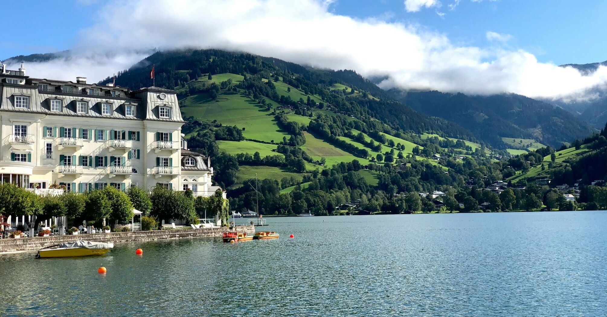 Scenic view of Zell am See with a historic lakeside hotel, green hills, and mist-covered mountains in the background