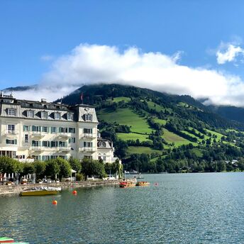 Scenic view of Zell am See with a historic lakeside hotel, green hills, and mist-covered mountains in the background