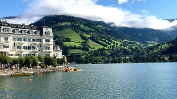 Scenic view of Zell am See with a historic lakeside hotel, green hills, and mist-covered mountains in the background