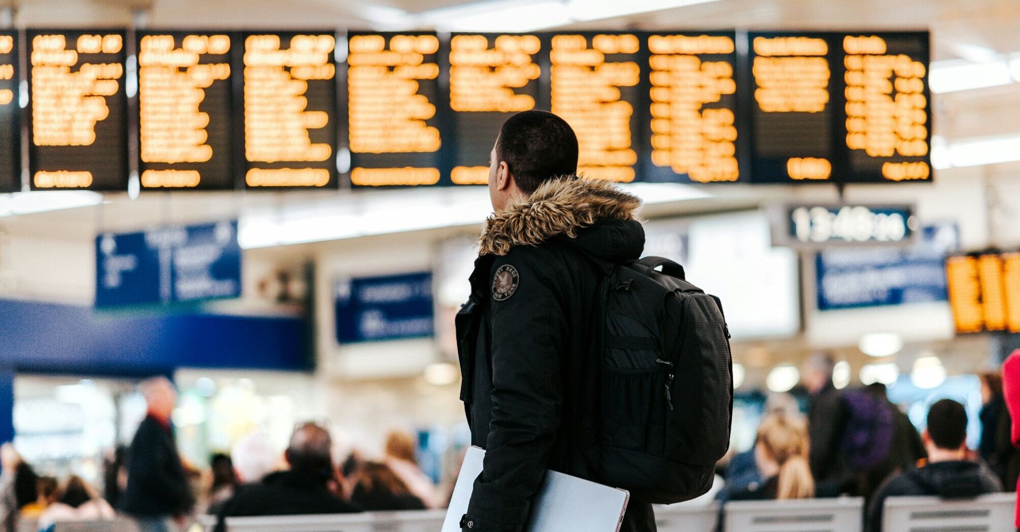 A traveler in a dark jacket with a backpack and laptop looks at the airport departure board, surrounded by other passengers