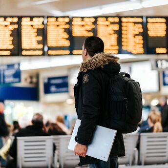 A traveler in a dark jacket with a backpack and laptop looks at the airport departure board, surrounded by other passengers
