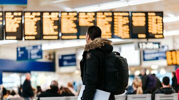 A traveler in a dark jacket with a backpack and laptop looks at the airport departure board, surrounded by other passengers