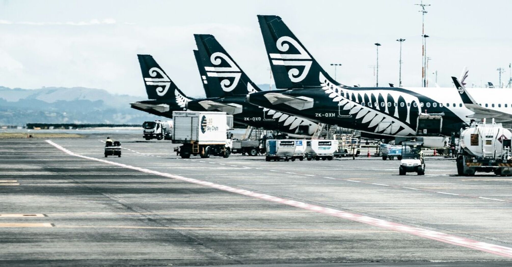 A row of Air New Zealand aircraft parked at an airport tarmac, with ground service vehicles operating nearby under a partly cloudy sky