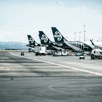 A row of Air New Zealand aircraft parked at an airport tarmac, with ground service vehicles operating nearby under a partly cloudy sky