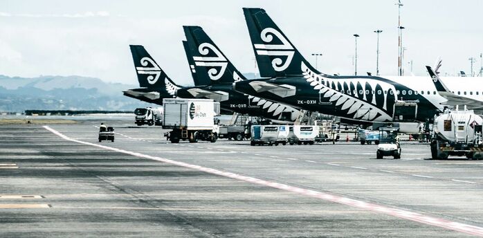 A row of Air New Zealand aircraft parked at an airport tarmac, with ground service vehicles operating nearby under a partly cloudy sky