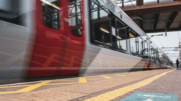 A red train in motion at a station platform with directional arrows on the ground