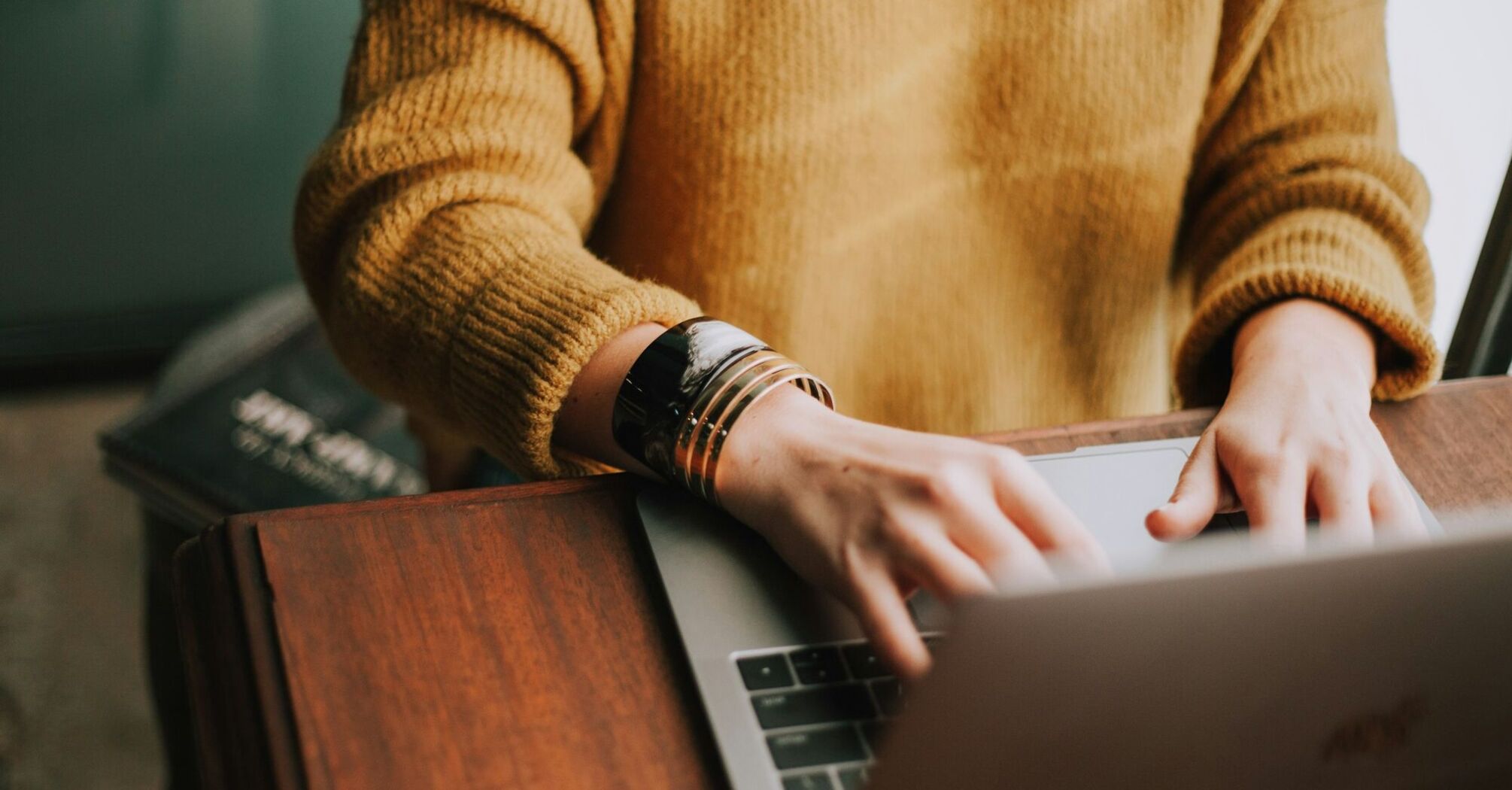 A person wearing a mustard-colored sweater typing on a laptop at a wooden desk