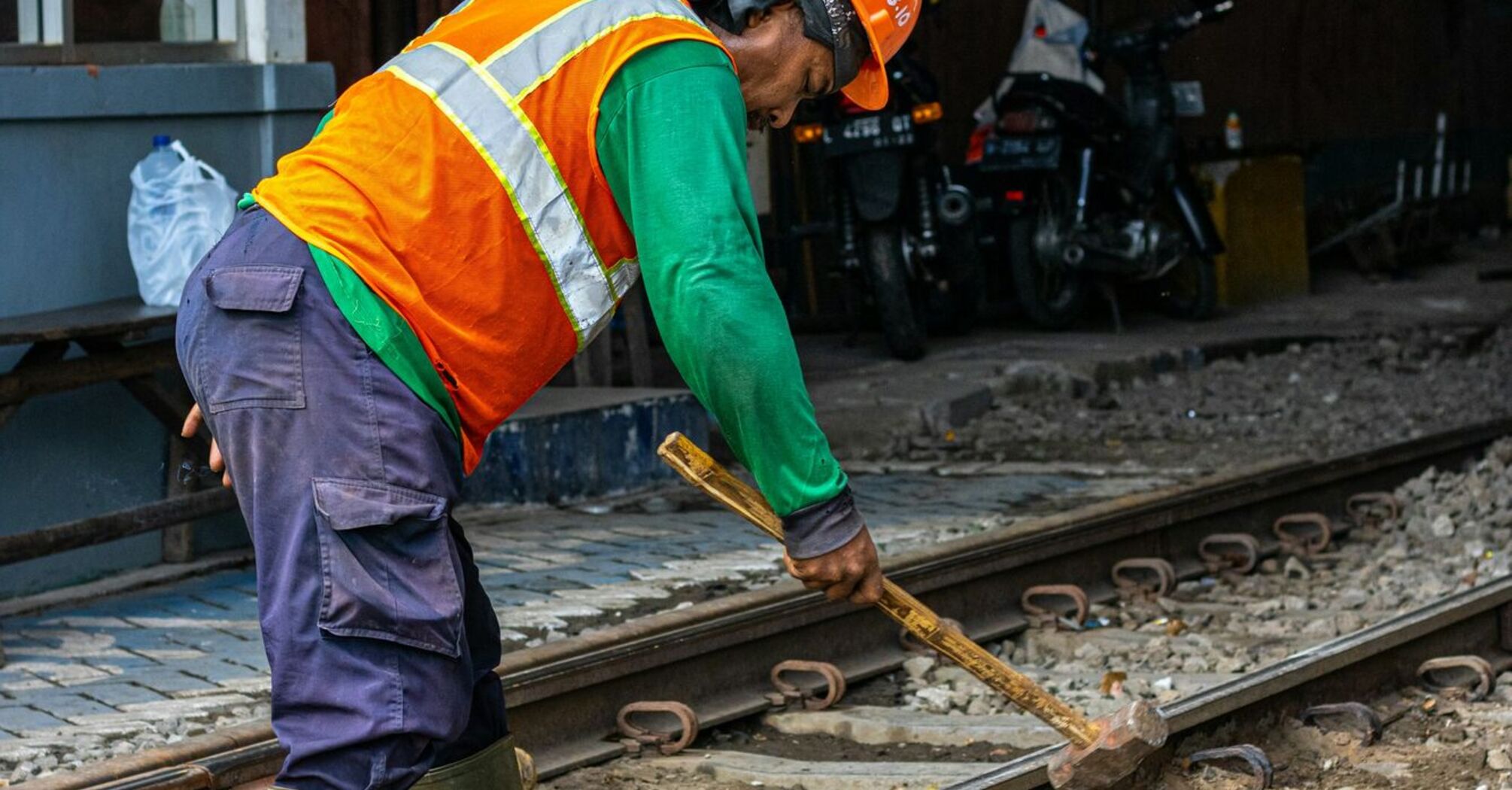 A railway worker in an orange safety vest and helmet repairing train tracks with a sledgehammer