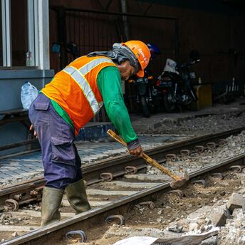 A railway worker in an orange safety vest and helmet repairing train tracks with a sledgehammer