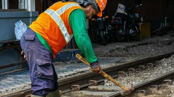 A railway worker in an orange safety vest and helmet repairing train tracks with a sledgehammer