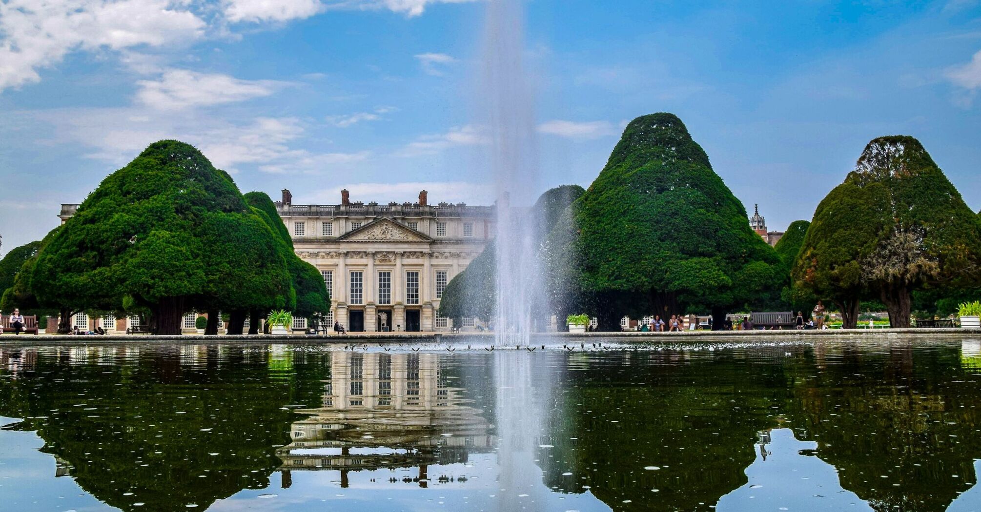 A scenic view of Hampton Court Palace's gardens with a fountain reflecting in the water