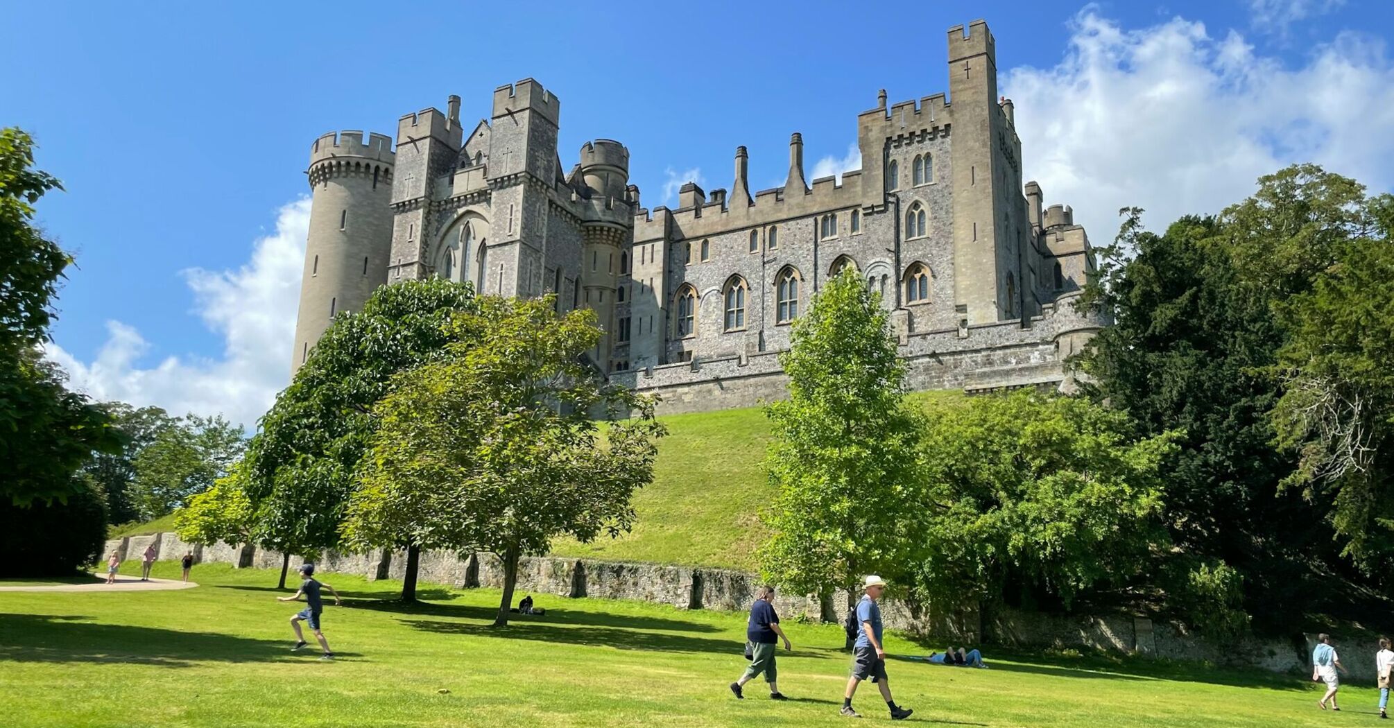 A view of Arundel Castle in West Sussex, surrounded by green lawns and trees, with visitors walking and enjoying the sunny day