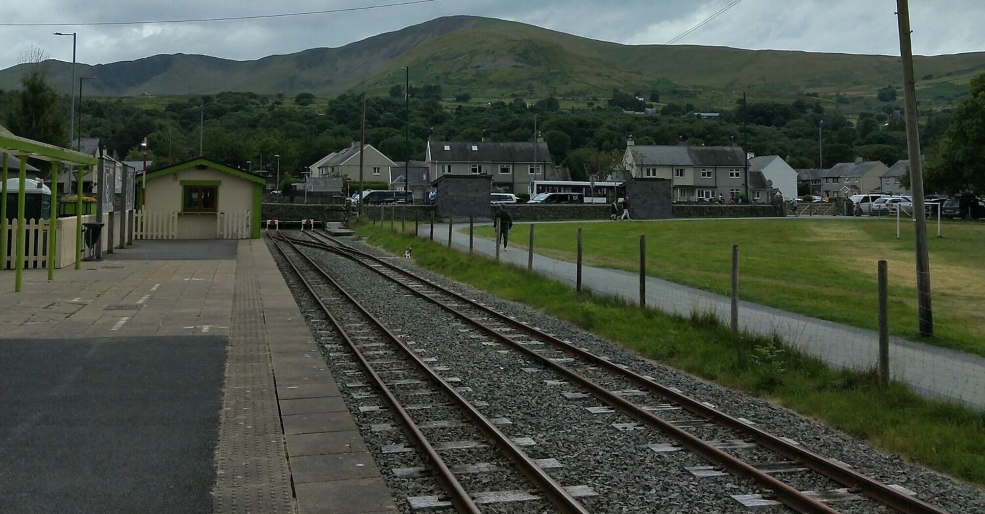 A quiet rural train station with railway tracks leading into the distance, set against green hills under a cloudy sky