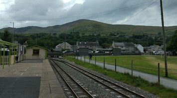 A quiet rural train station with railway tracks leading into the distance, set against green hills under a cloudy sky