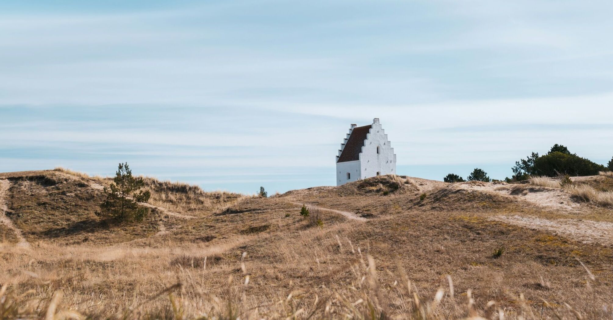 A white church partially buried in sand dunes under a vast blue sky in Jutland, Denmark