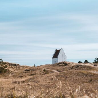 A white church partially buried in sand dunes under a vast blue sky in Jutland, Denmark