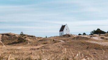 A white church partially buried in sand dunes under a vast blue sky in Jutland, Denmark