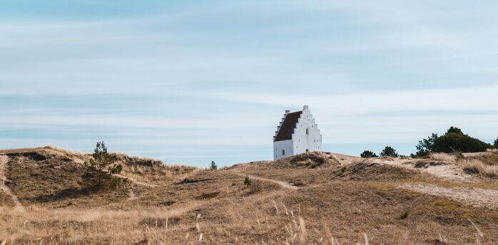 A white church partially buried in sand dunes under a vast blue sky in Jutland, Denmark