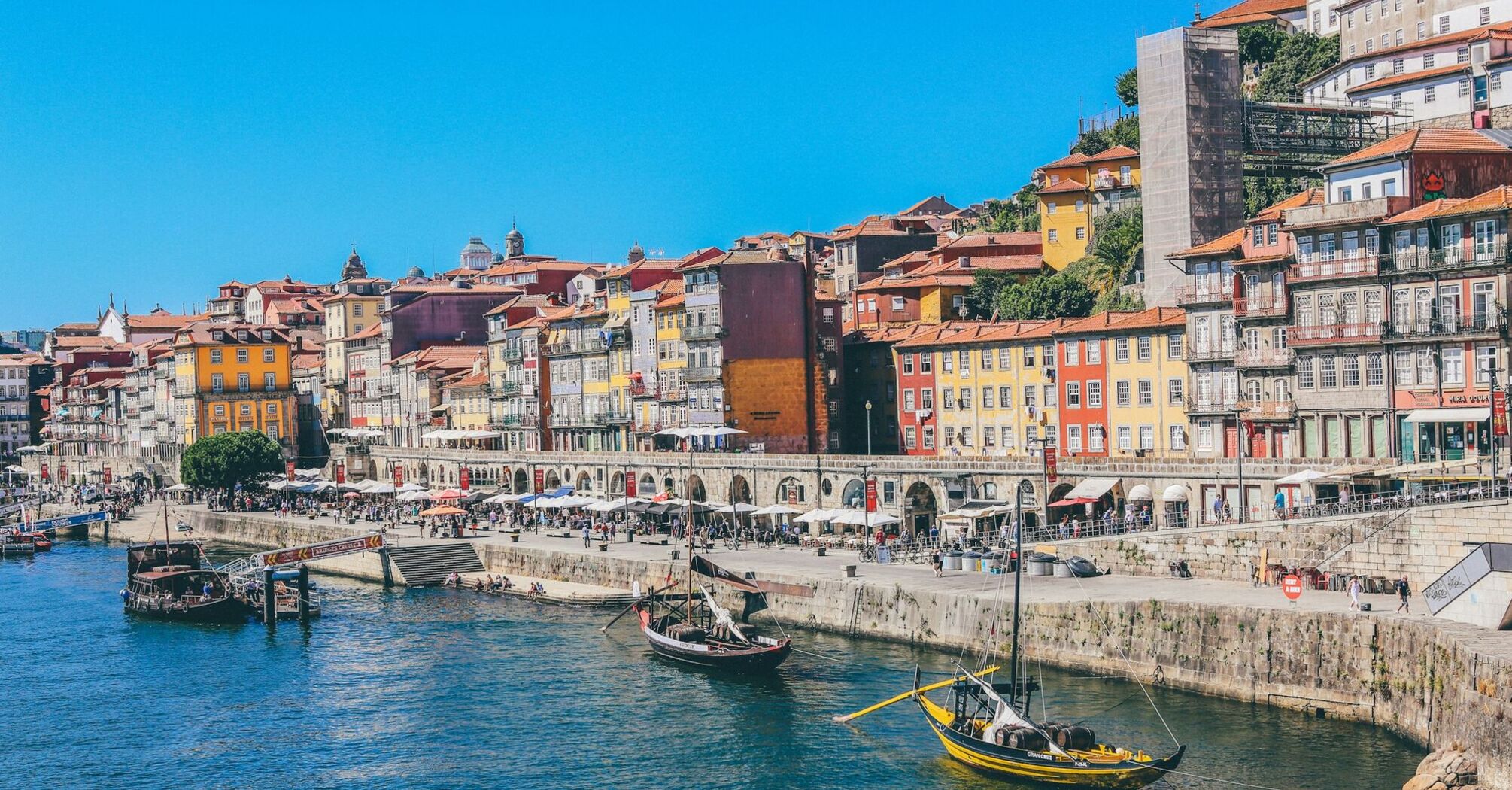 Colorful historic buildings line the waterfront of Porto, Portugal, with traditional boats floating on the Douro River under a bright blue sky