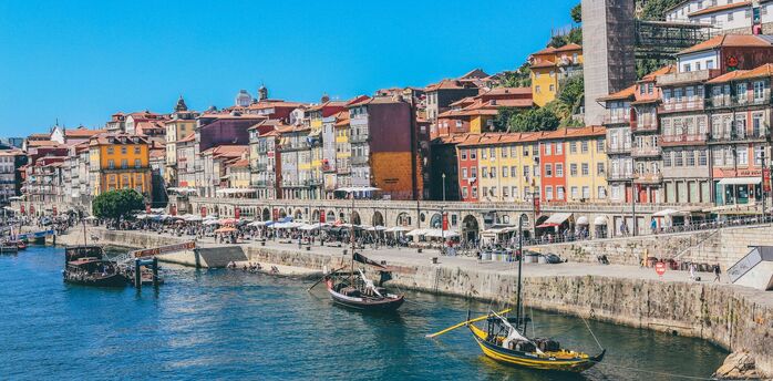 Colorful historic buildings line the waterfront of Porto, Portugal, with traditional boats floating on the Douro River under a bright blue sky