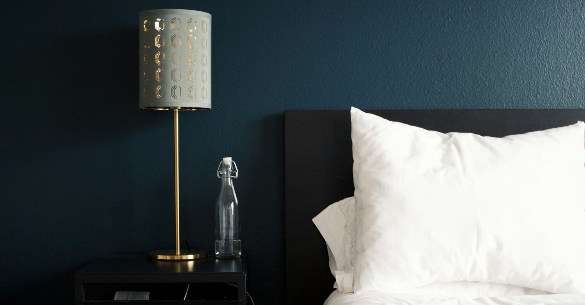 A modern hotel room with a black nightstand, a gold lamp, a glass water bottle, and a neatly arranged white pillow on a dark bed frame