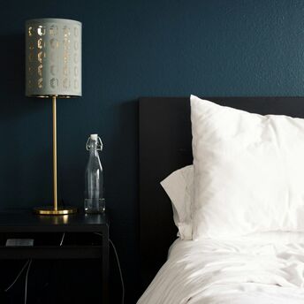 A modern hotel room with a black nightstand, a gold lamp, a glass water bottle, and a neatly arranged white pillow on a dark bed frame