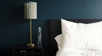 A modern hotel room with a black nightstand, a gold lamp, a glass water bottle, and a neatly arranged white pillow on a dark bed frame