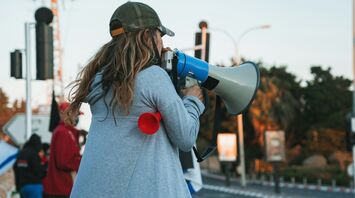 A woman in a gray hoodie and a green cap speaks into a megaphone during a protest, with other demonstrators visible in the background