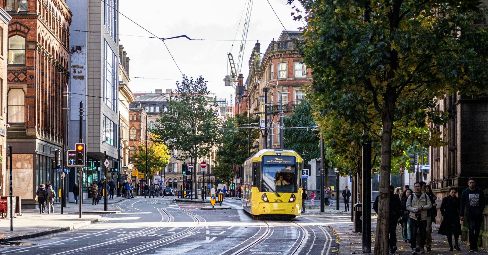 A yellow tram travels through a busy street in Manchester, surrounded by pedestrians and historic buildings