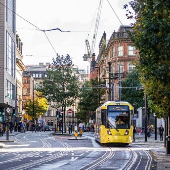 A yellow tram travels through a busy street in Manchester, surrounded by pedestrians and historic buildings